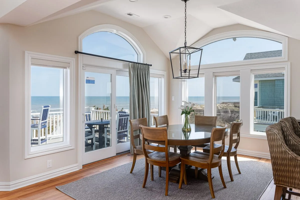 Comedor con mesa de madera y asientos junto a grandes ventanas con vista al mar.