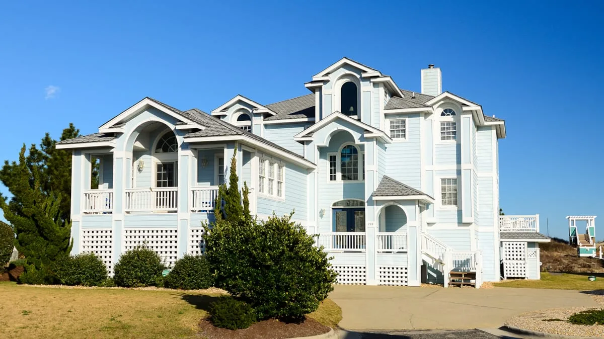 A large coastal house with white siding, porches, and shrubs in the yard.