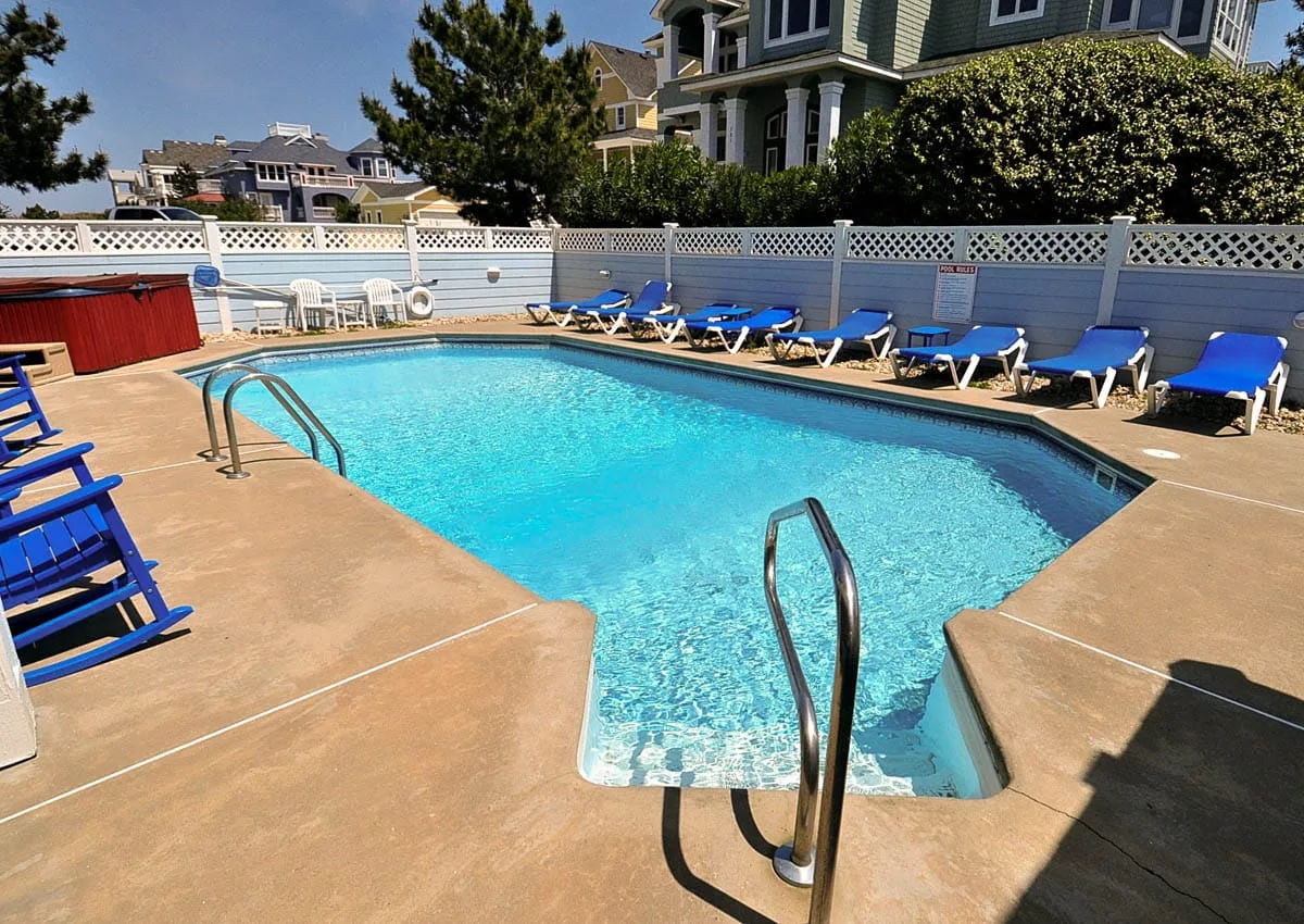 An outdoor swimming pool is surrounded by lounge chairs and a white fence.
