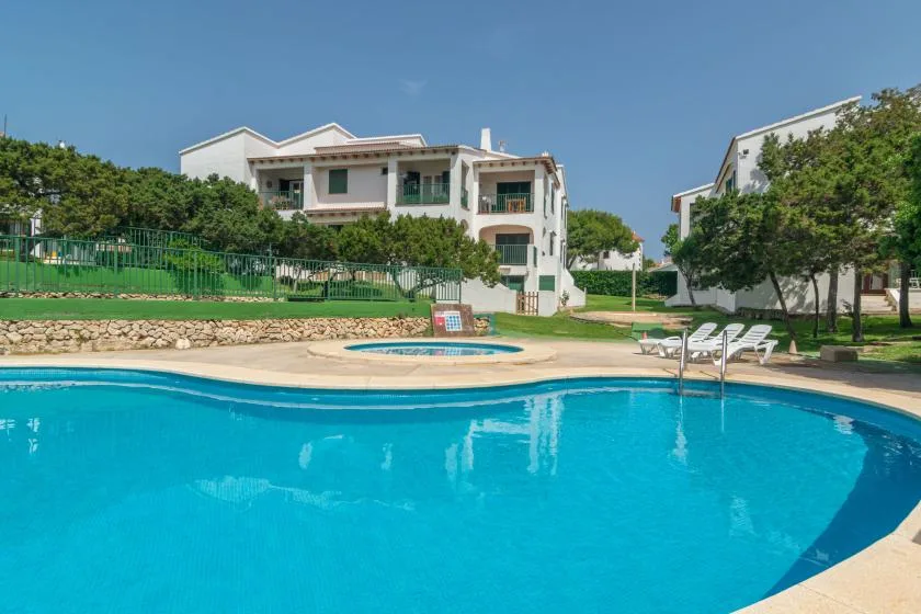 Piscina al aire libre junto a edificios blancos y jardines bajo un cielo despejado.