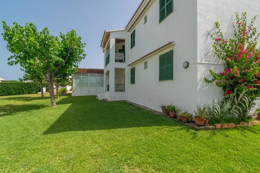 Una casa blanca de varias plantas da a un césped verde con árboles y plantas con flores.