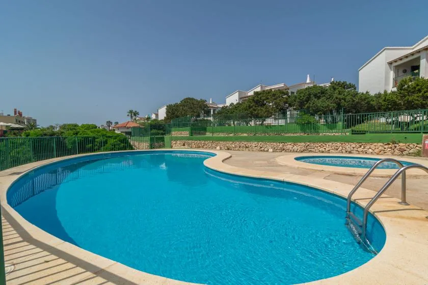 Una piscina al aire libre está junto a una piscina más pequeña, con edificios y vegetación al fondo.