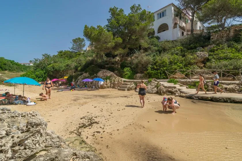 Una playa de arena está bajo una colina verde, con personas caminando por la orilla.