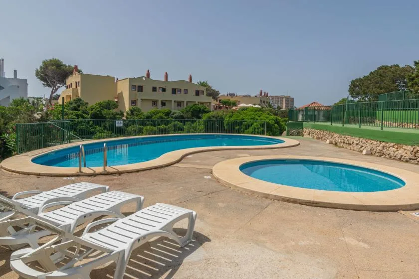 Piscinas al aire libre junto a una terraza pavimentada con tumbonas blancas.
