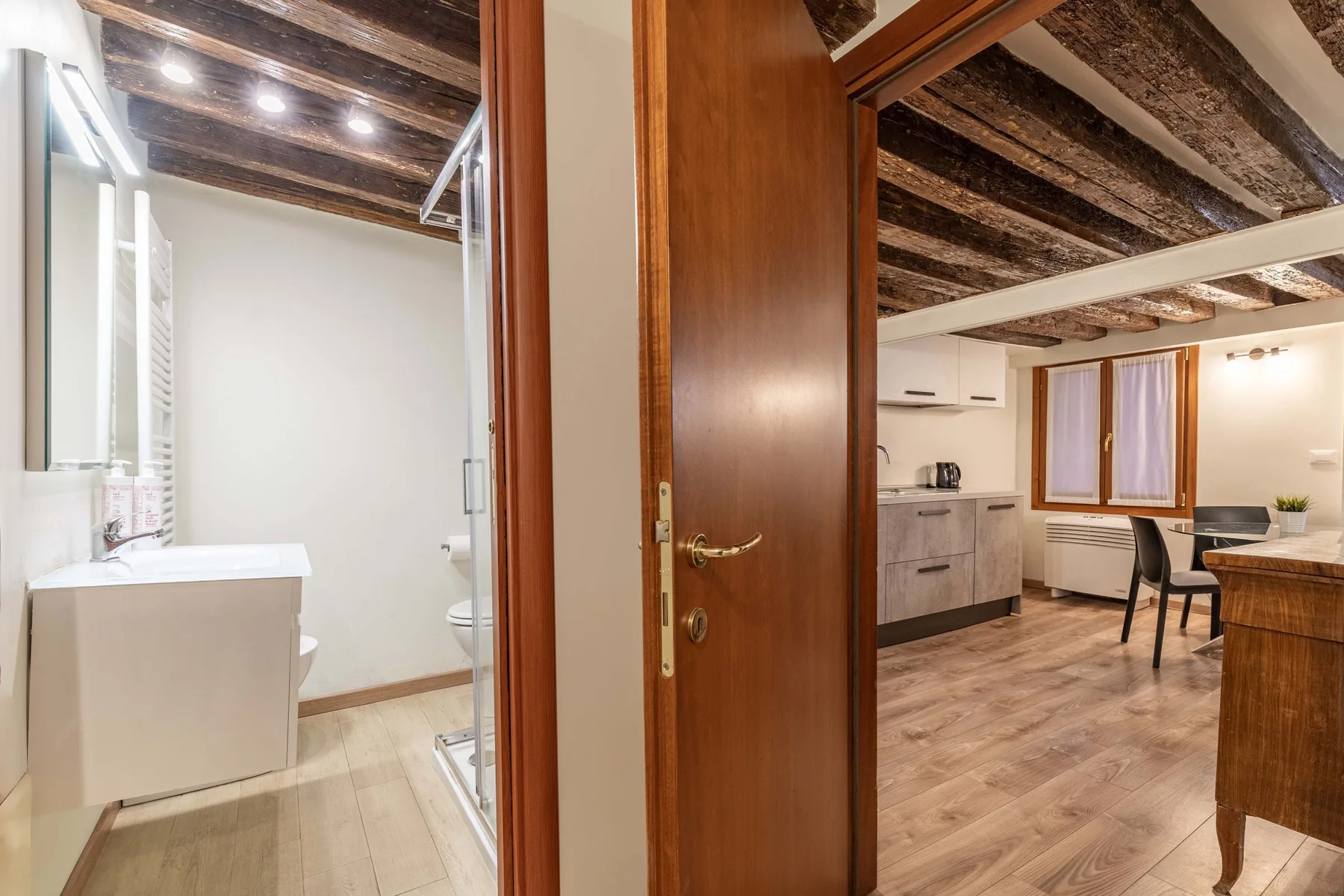 Una puerta de madera abierta conecta un baño con una zona de comedor y cocina.