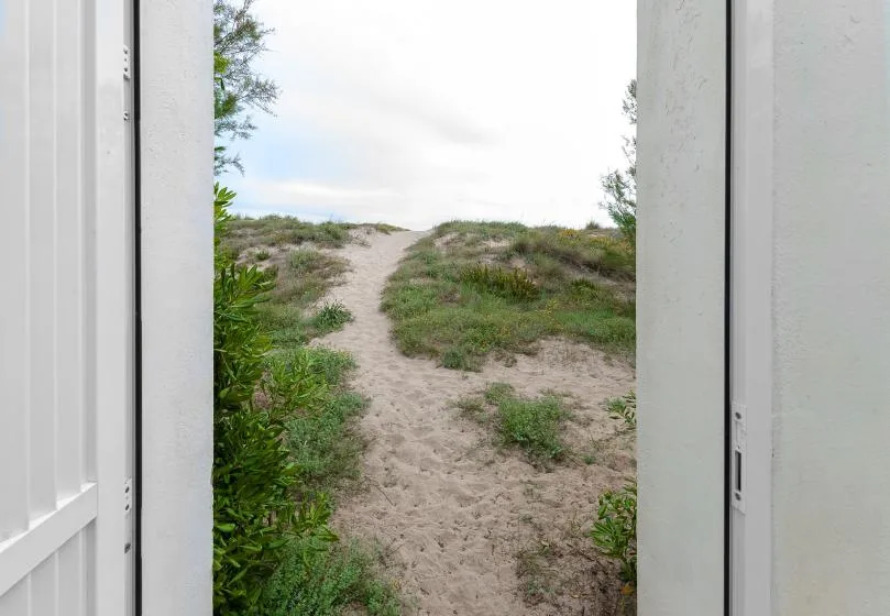 A sandy path winds through low vegetation toward the beach, viewed from a doorway.