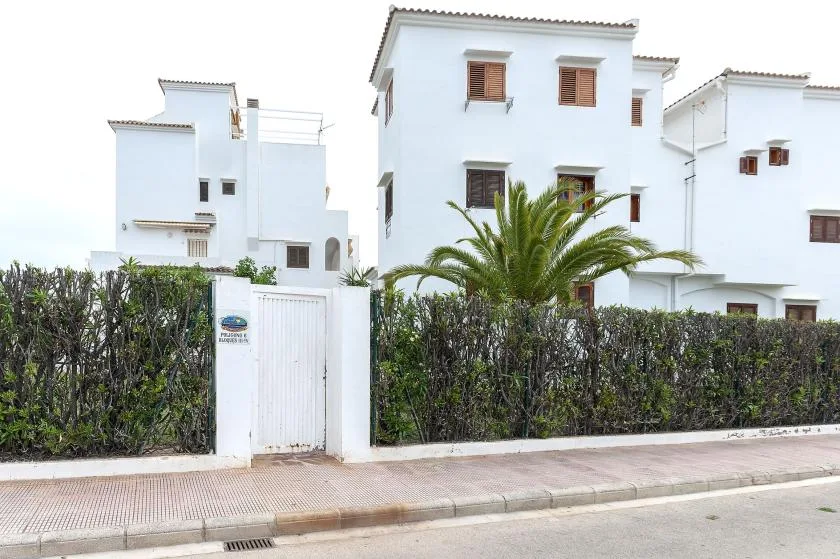 White buildings stand behind a hedge along a paved sidewalk.