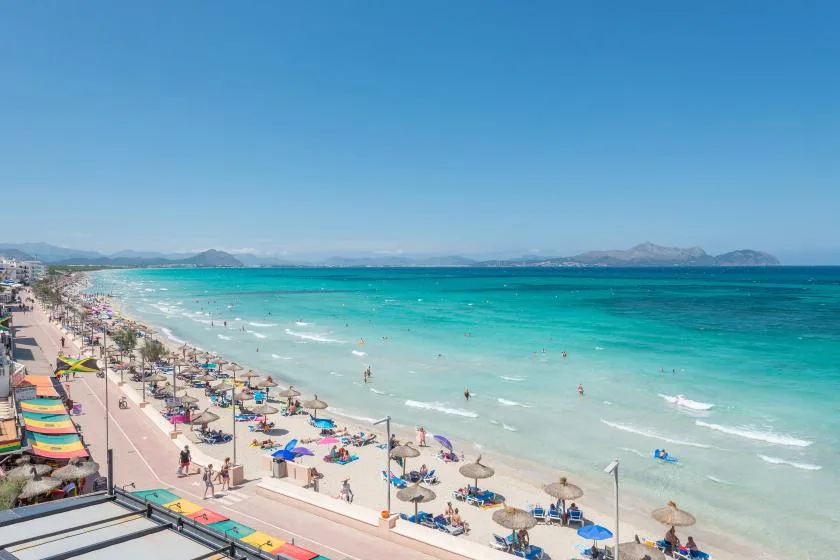 A busy sandy beach stretches along bright turquoise water beneath a clear blue sky.