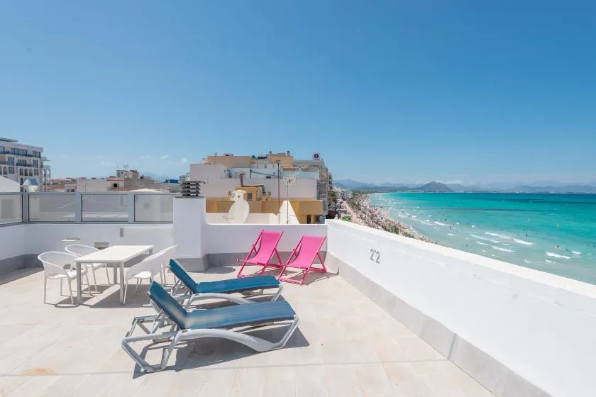 A seaside rooftop terrace with lounge seating, a dining table, and bright turquoise water.