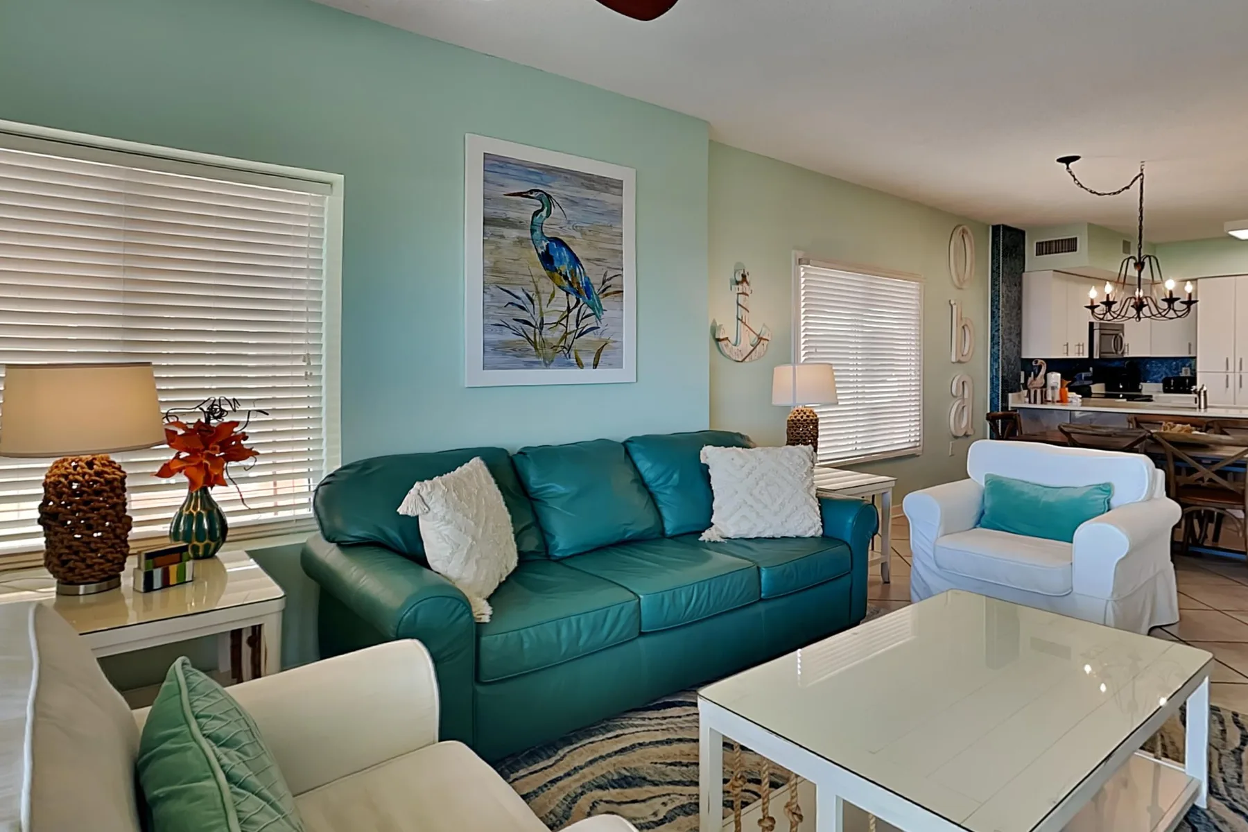 A living room with teal seating, a white coffee table, and windows with blinds.