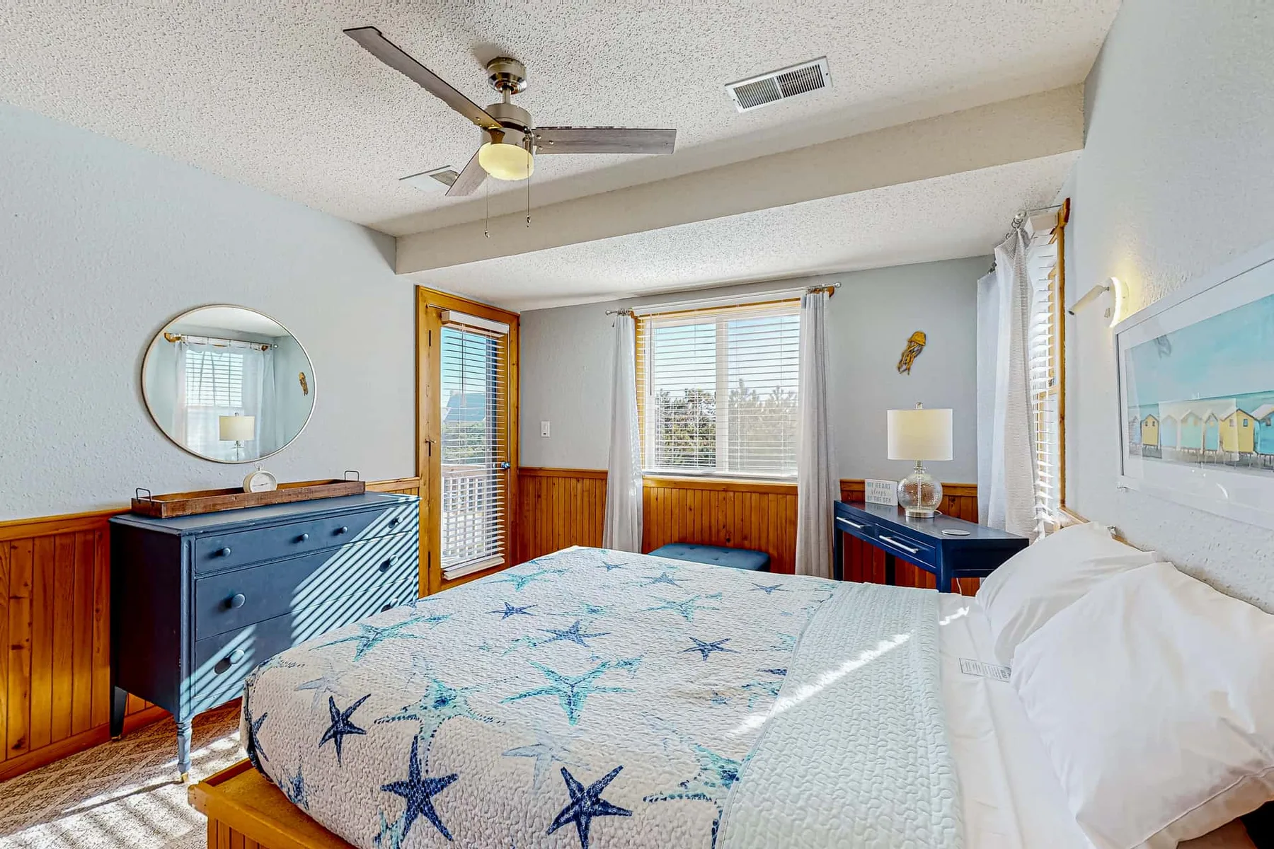 A bedroom with a star-patterned bedspread, wooden wall paneling, and windows.