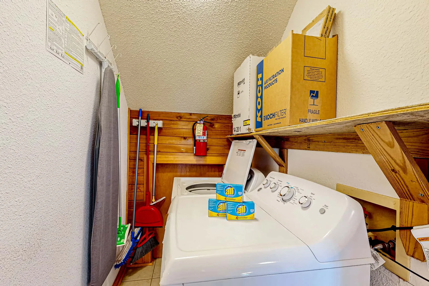 A laundry area with a washing machine and detergent beneath a wooden shelf.