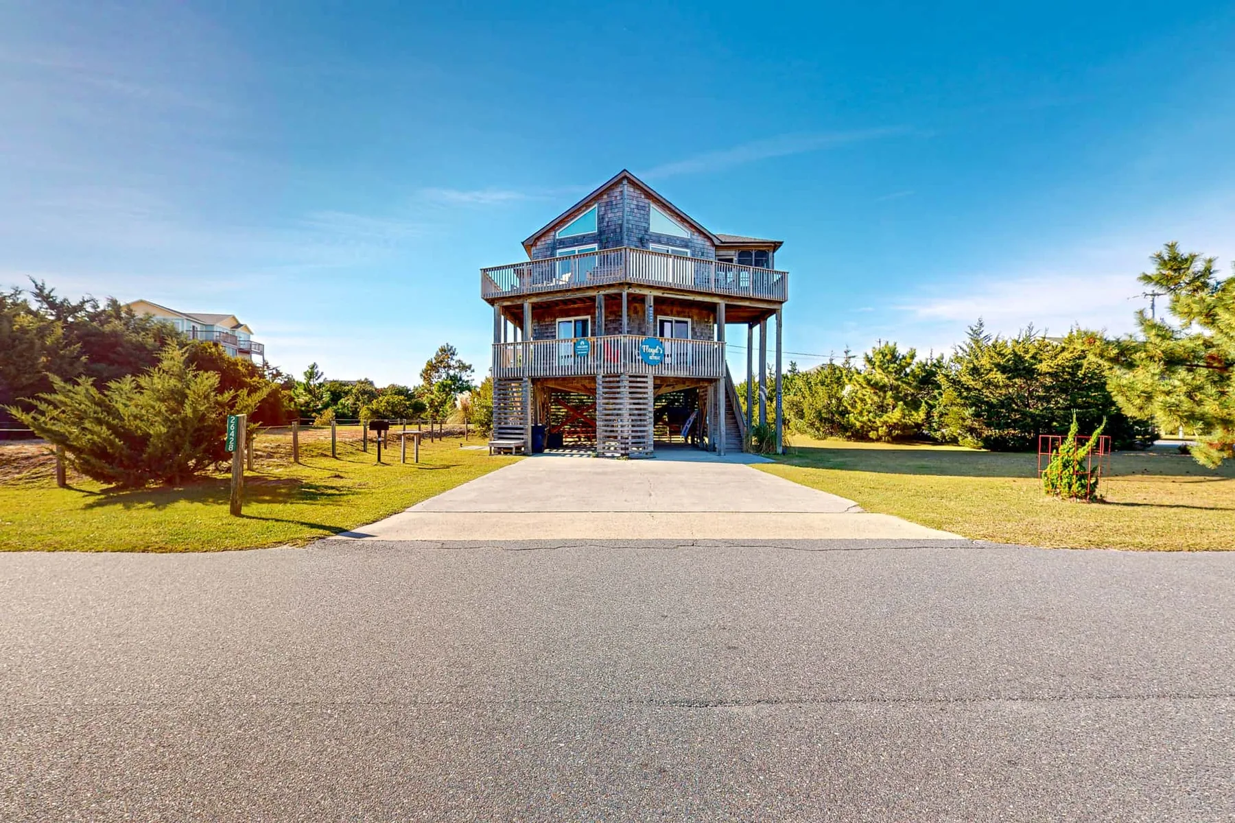 A raised seaside-style house stands beyond a paved driveway, surrounded by trees.