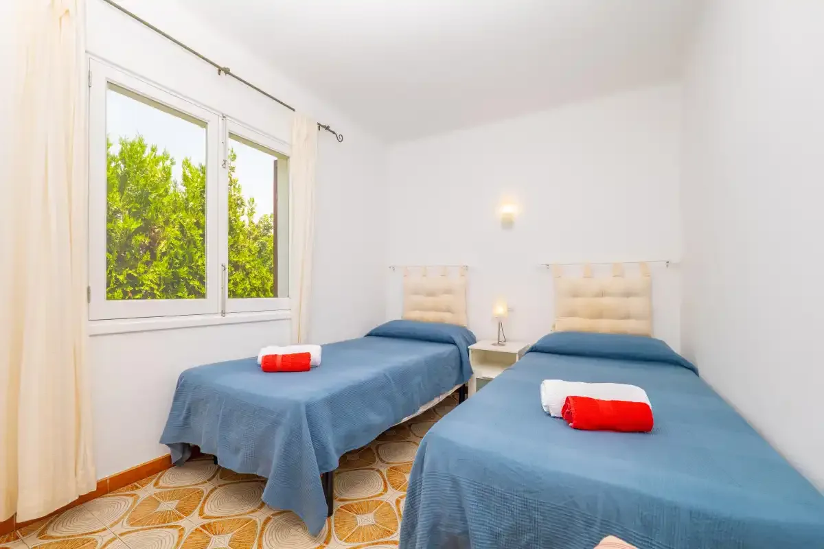A bright bedroom with two beds covered in blue bedspreads and a large window.