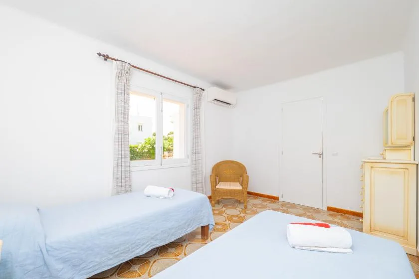 A bright bedroom with two beds, a window, and a woven chair.