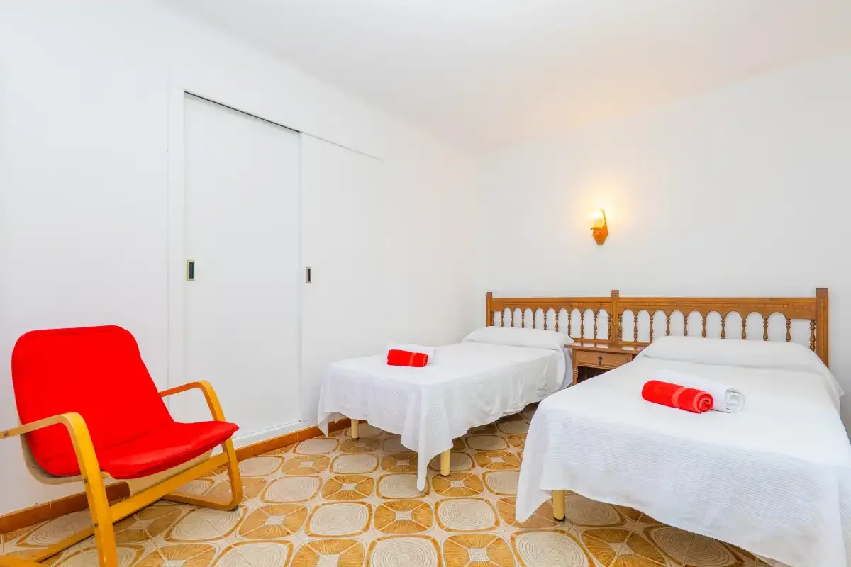 A bright bedroom with two beds and a red lounge chair.