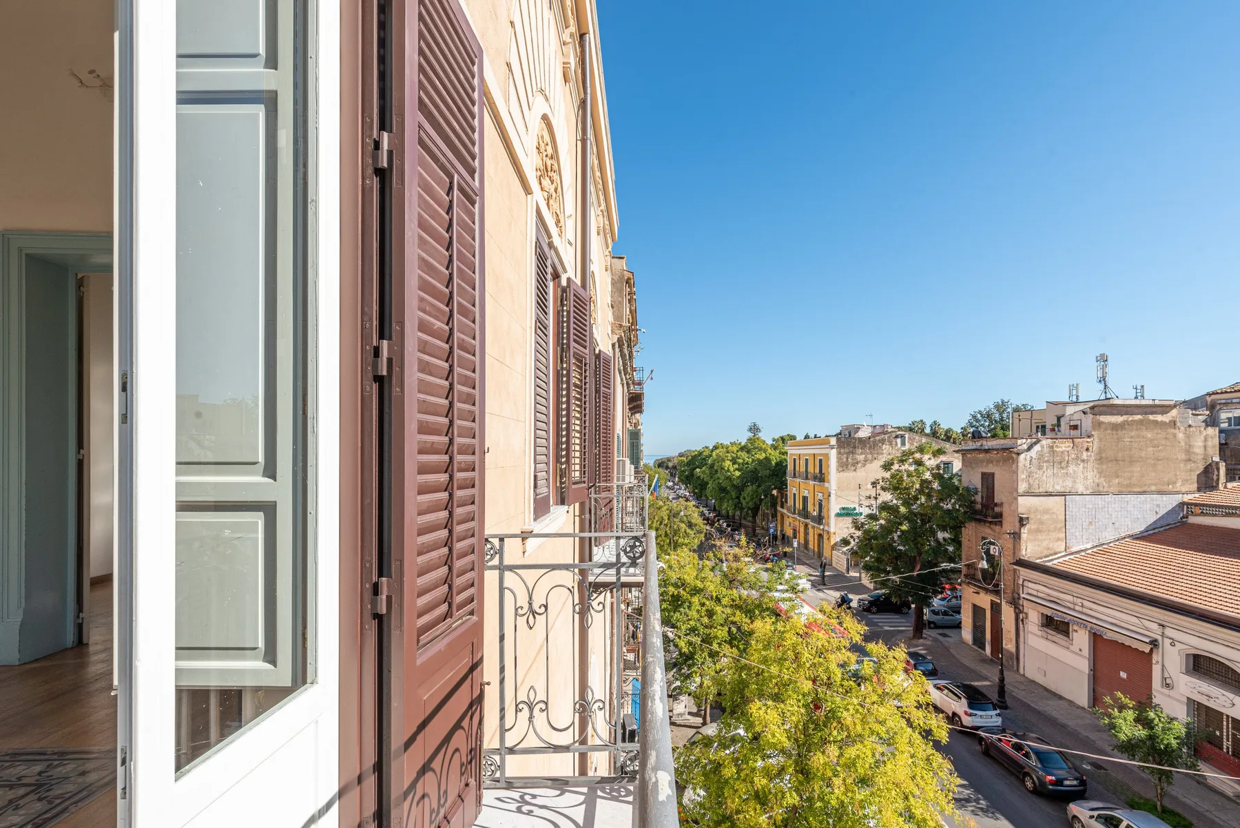 Un balcón da a una calle urbana soleada, bordeada de edificios y árboles.