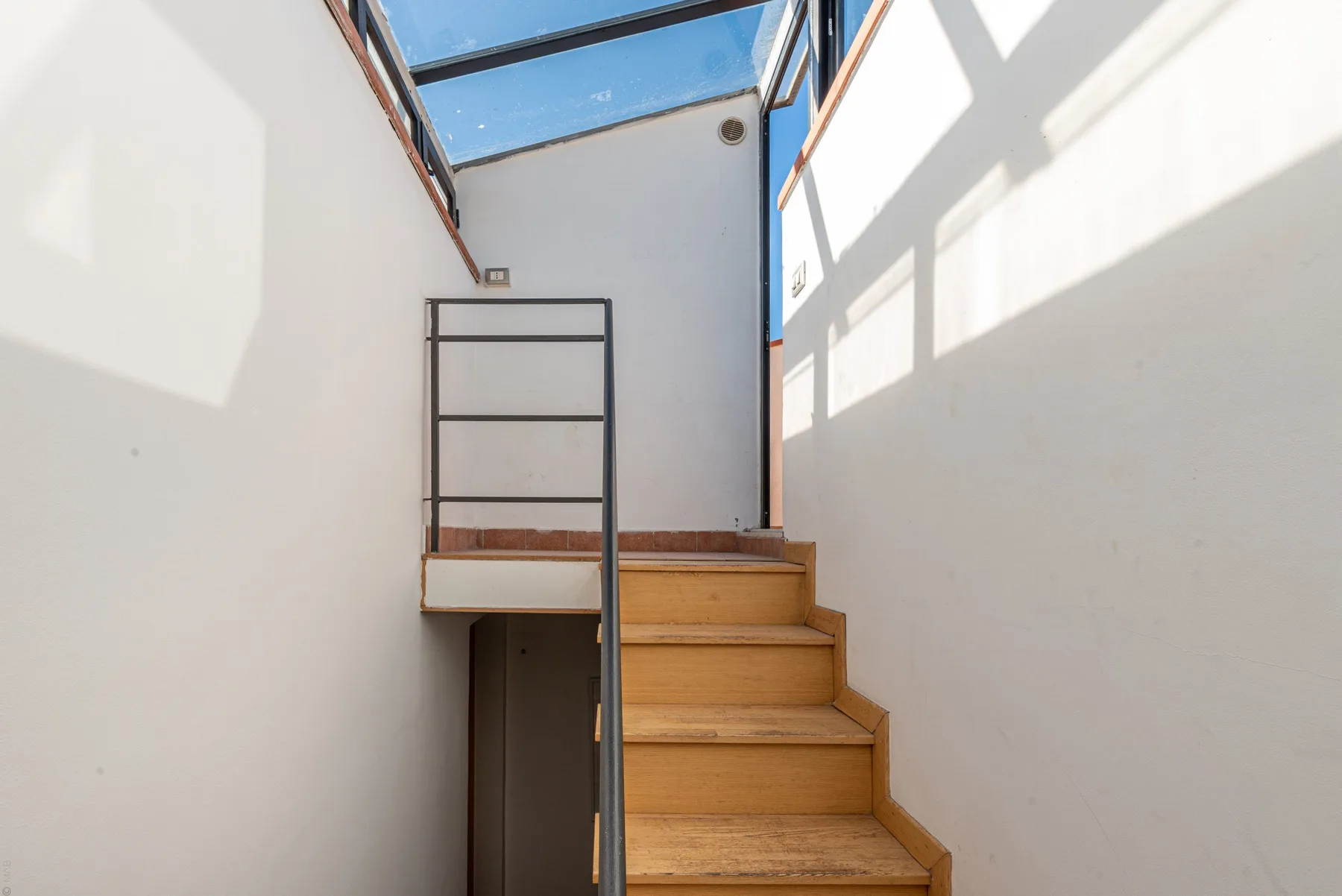 Una escalera de madera sube bajo una claraboya de vidrio en un interior luminoso.