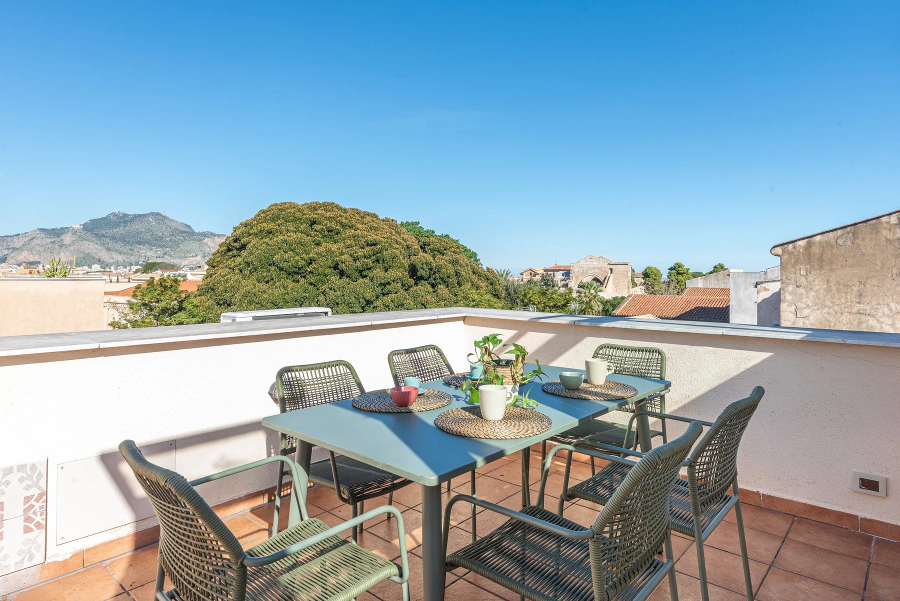 Una terraza en la azotea tiene una mesa de comedor y asientos con vistas a edificios y colinas cercanas.
