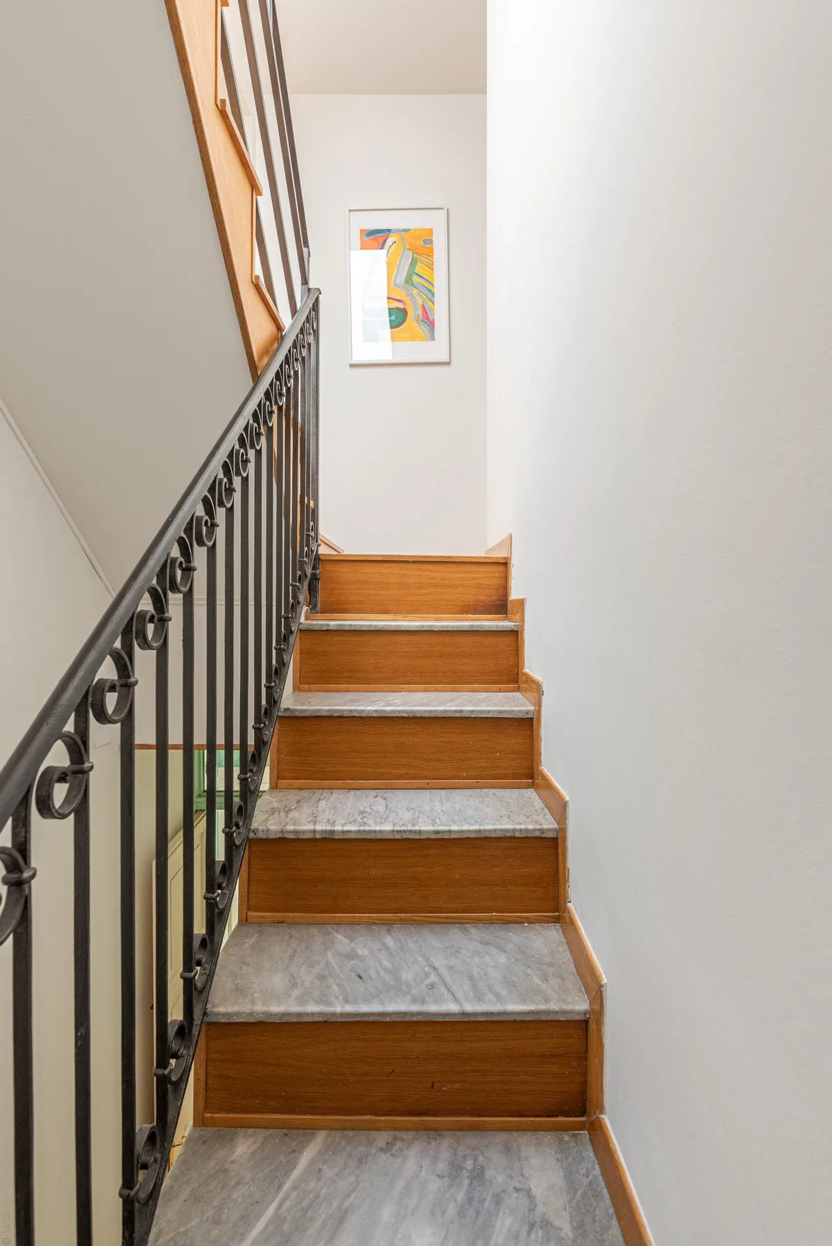 Una escalera con peldaños de madera y una barandilla oscura conduce a una obra enmarcada.