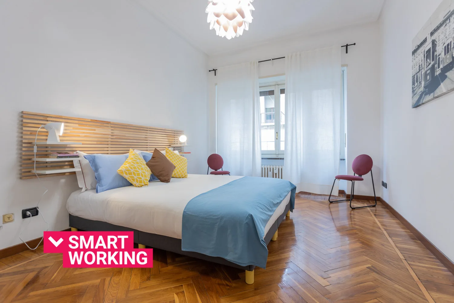 Un dormitorio luminoso con una cama, cojines de colores y ventanas altas con cortinas.