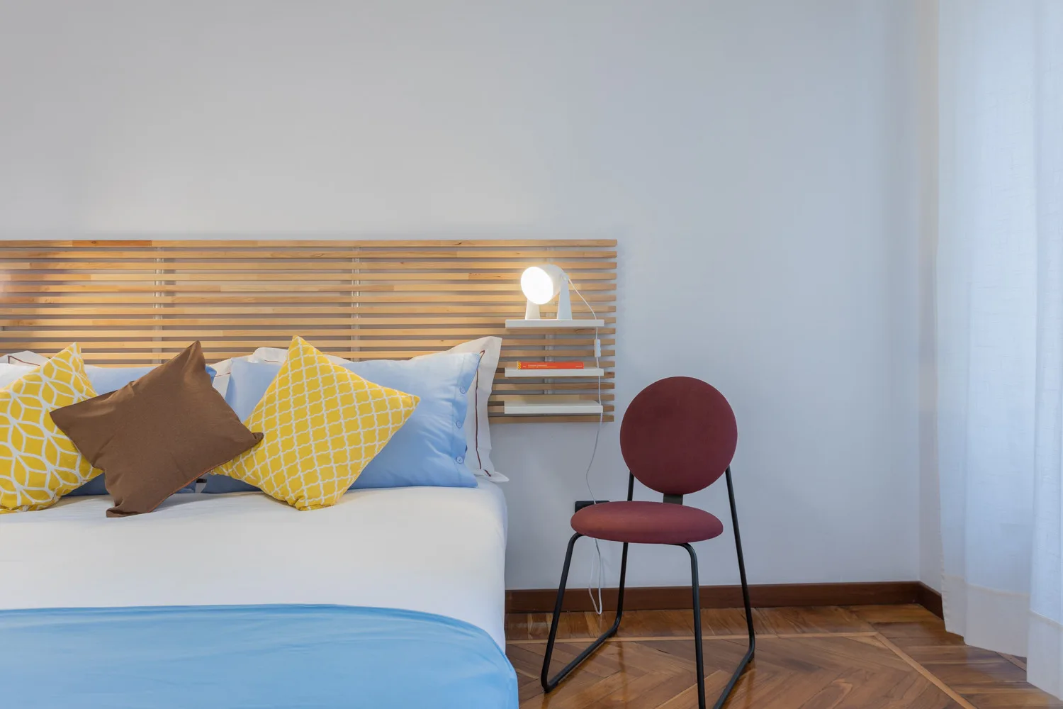 A bedroom with a bed, colorful pillows, and a red chair beside it.