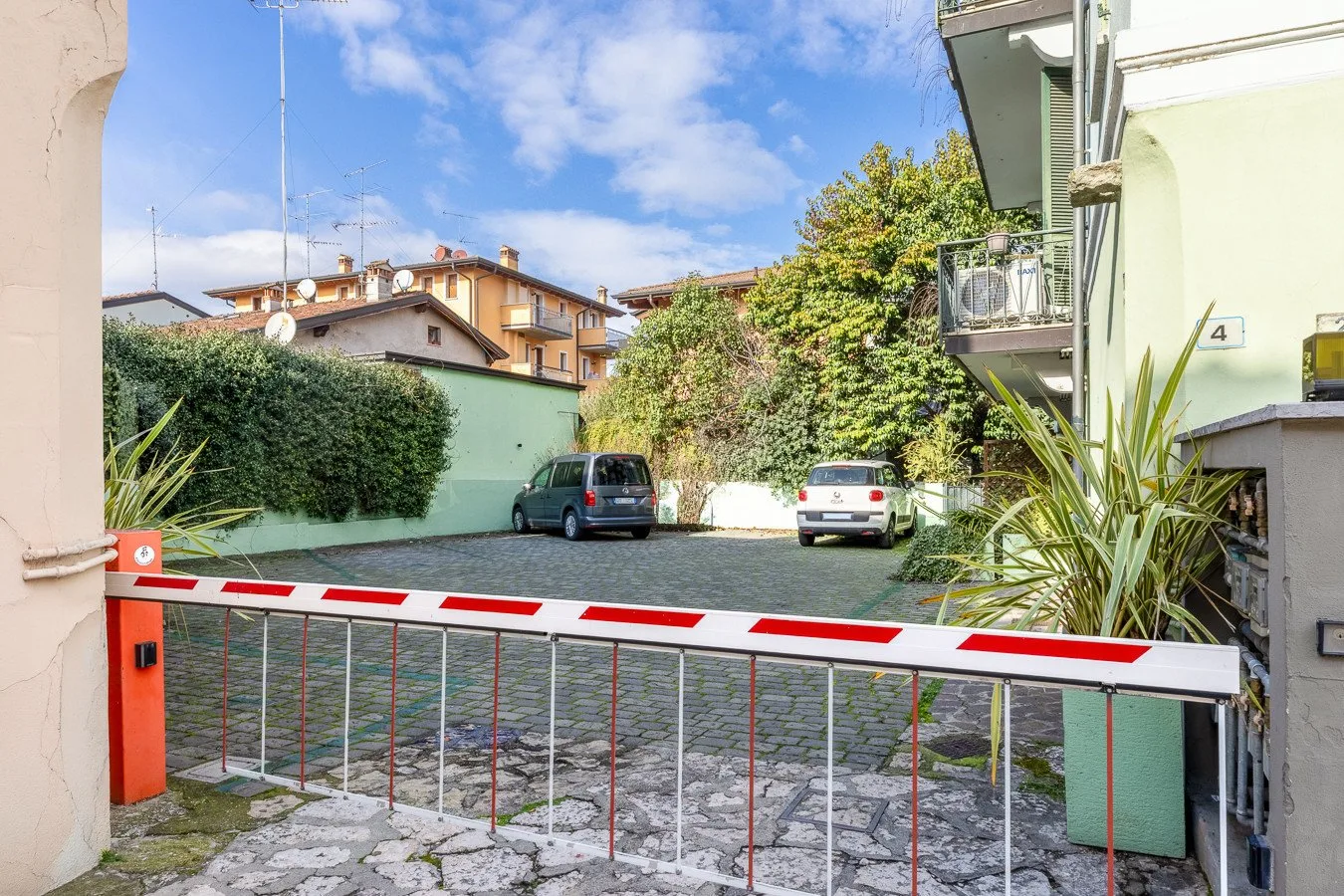 Una zona de estacionamiento pavimentada queda tras una barrera roja y blanca, con coches y edificios alrededor.