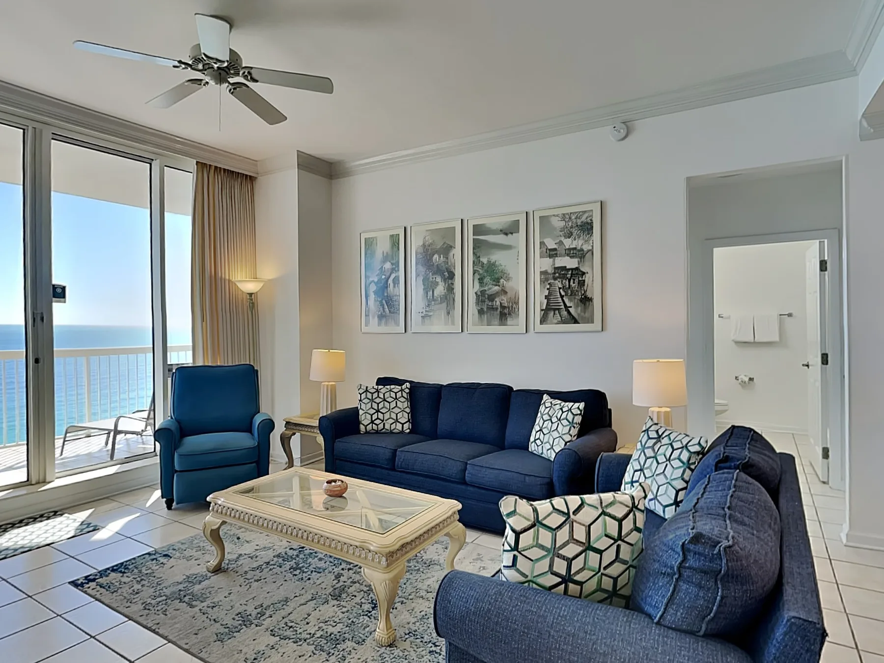 A living room with blue seating, a glass coffee table, and ocean views through the windows.