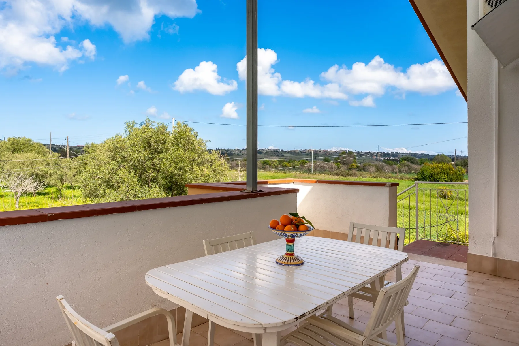 Una terraza exterior con suelo de baldosas tiene una mesa blanca con vistas a campos verdes.