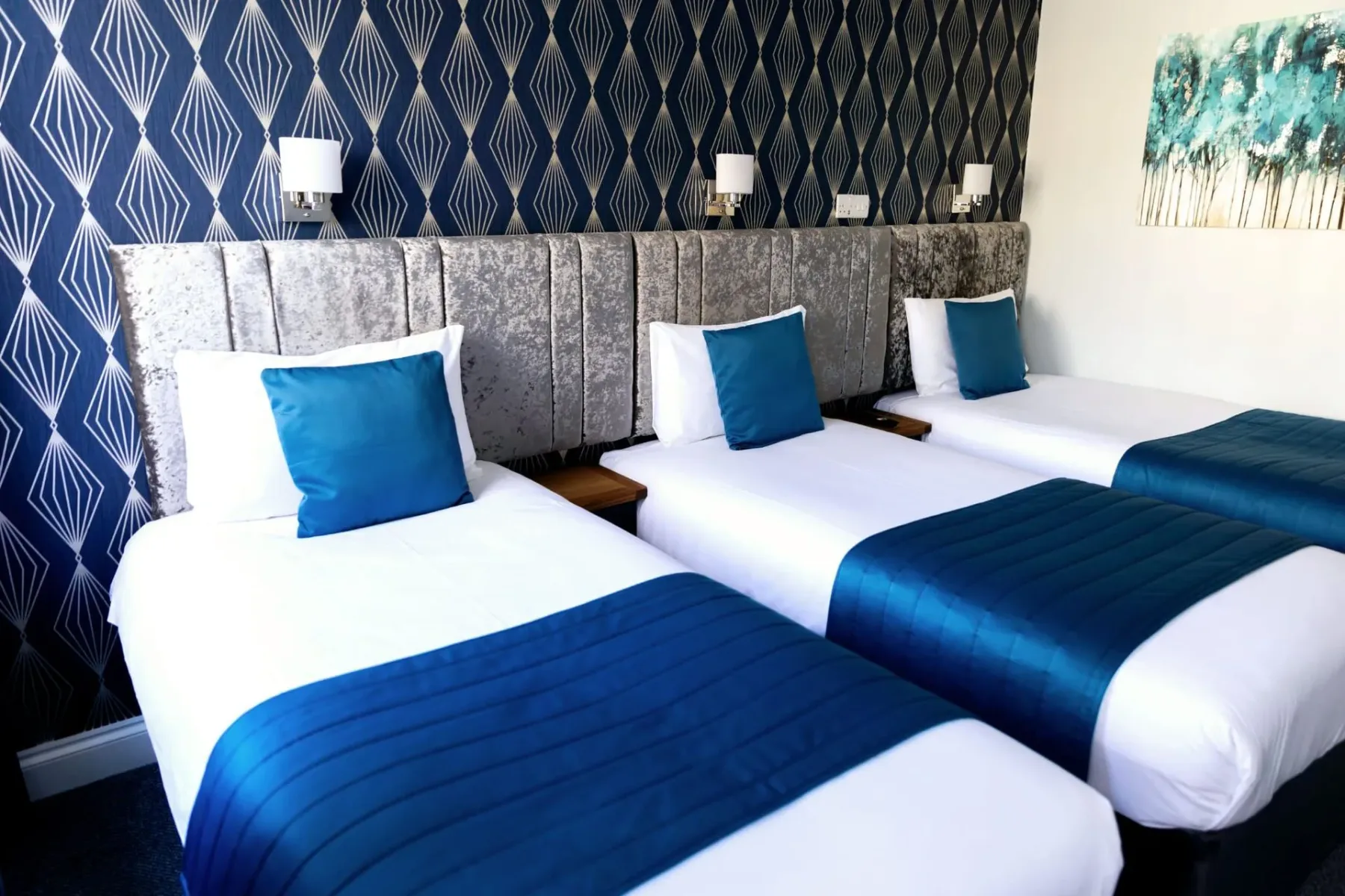 A hotel room with three beds and blue blankets.