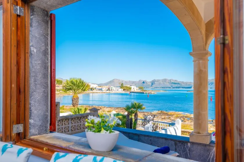 Una ventana arqueada abierta da a una bahía azul, con plantas en el alféizar.