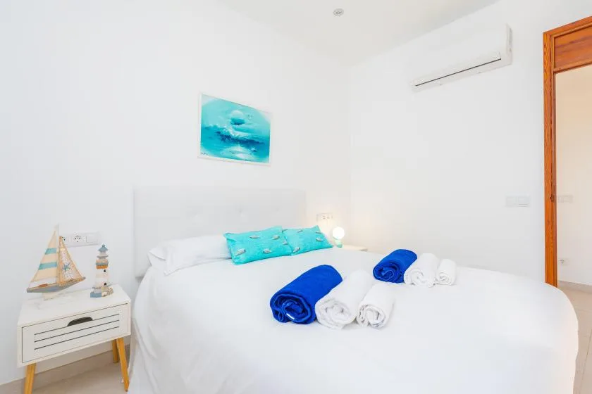 Un dormitorio luminoso con una cama blanca, cojines turquesa y toallas dobladas.