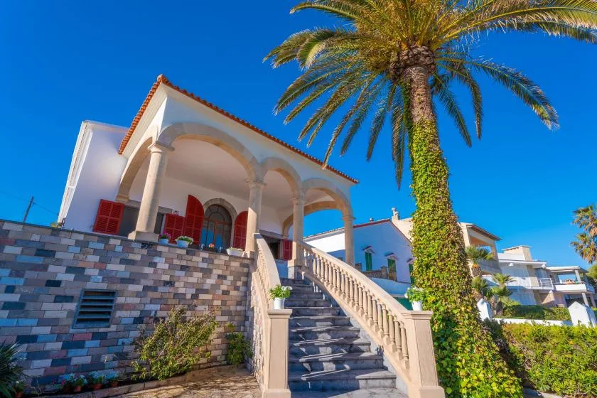 Un edificio de estilo mediterráneo con terraza de arcos, escaleras de piedra y una palmera.
