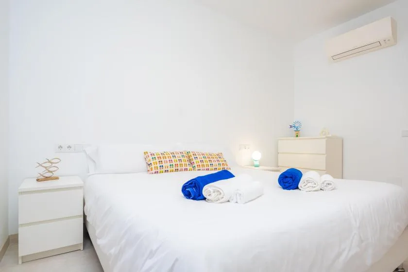Un dormitorio luminoso con ropa de cama blanca, almohadas coloridas y toallas dobladas sobre la cama.