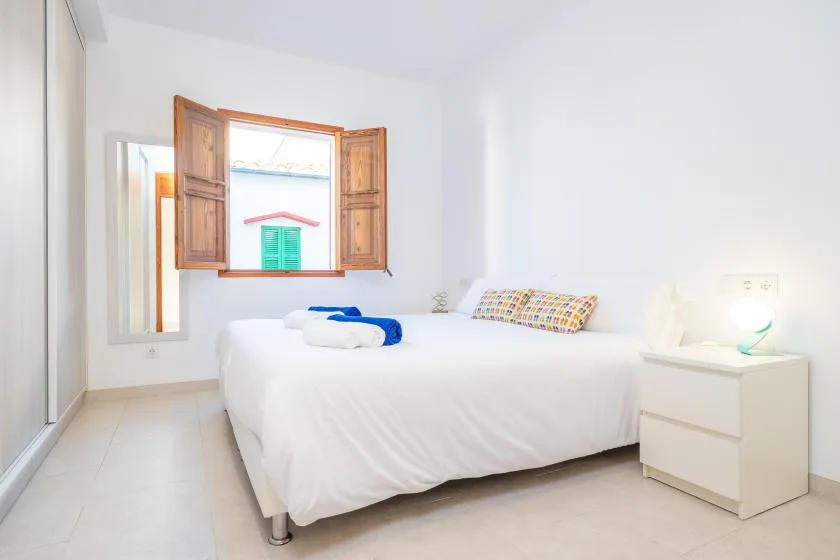Un dormitorio luminoso con una cama bajo una ventana con contraventanas de madera abiertas.