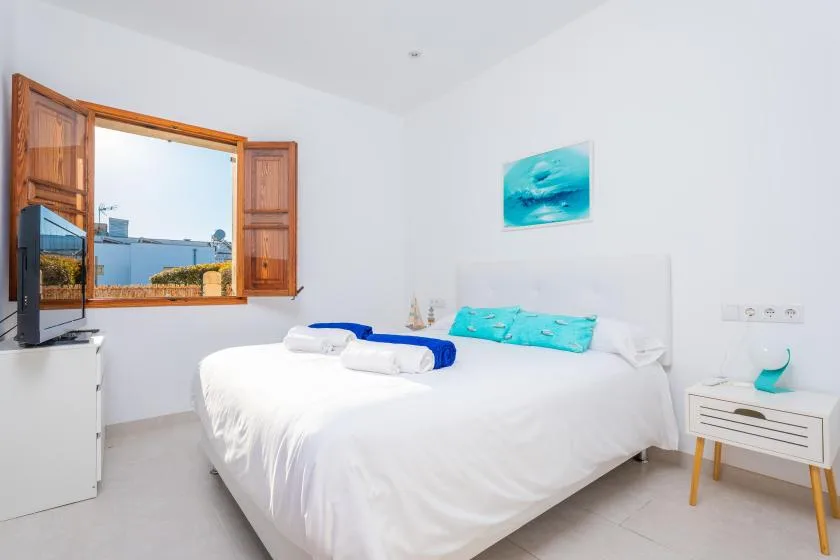 Un dormitorio luminoso con una cama blanca, cojines turquesa y una ventana abierta.
