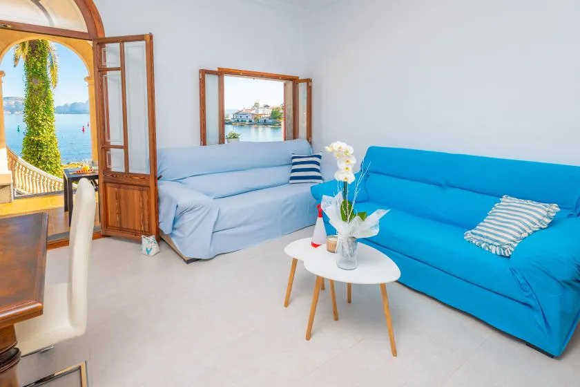 Una sala luminosa con asientos azules, una mesa pequeña y ventanas abiertas.