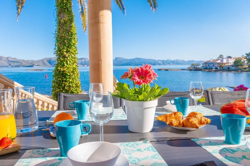 Una mesa junto al agua está preparada con tazas turquesas, bollería y flores.