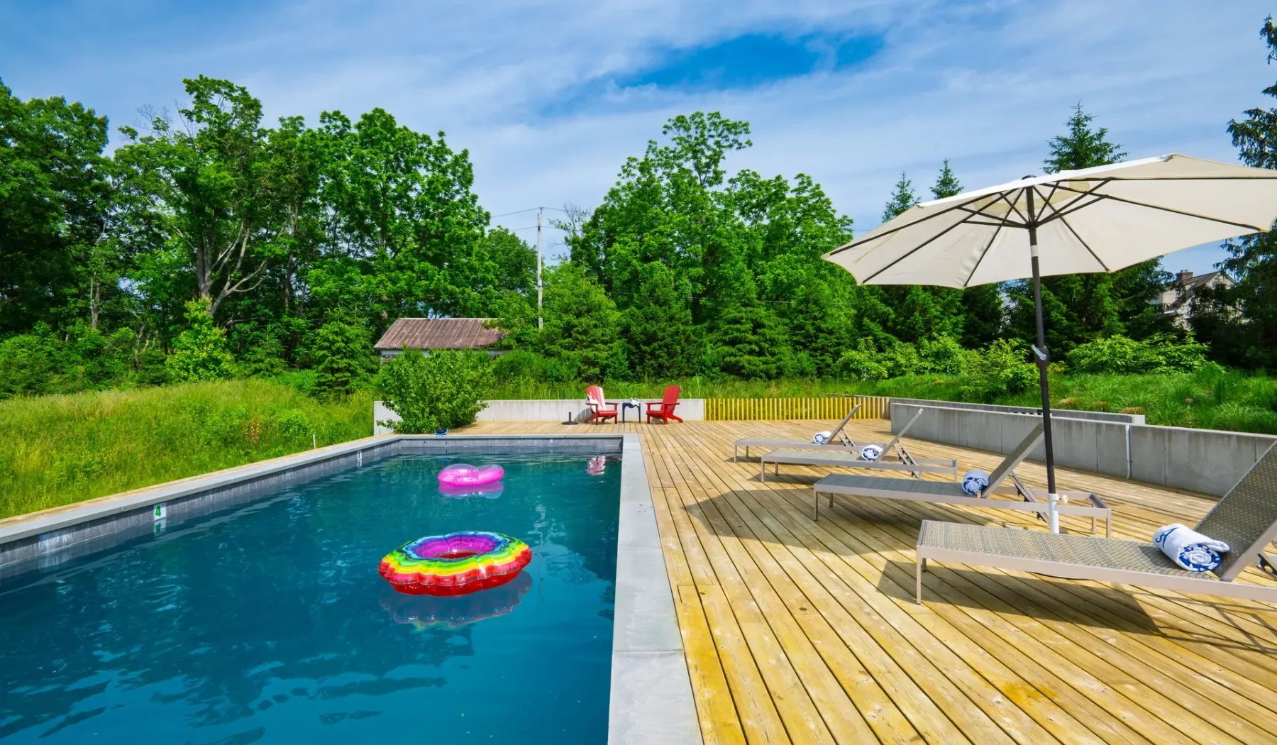 Una piscina al aire libre junto a una terraza de madera con asientos reclinables y una sombrilla.