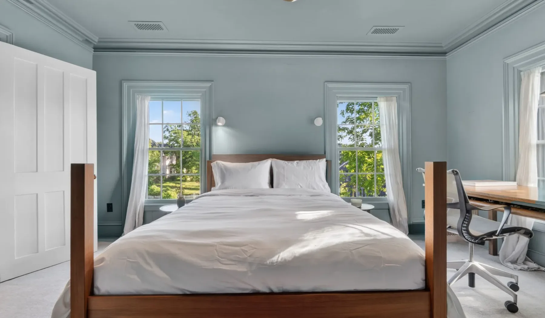 Un dormitorio con una cama entre dos ventanas y un escritorio cerca.