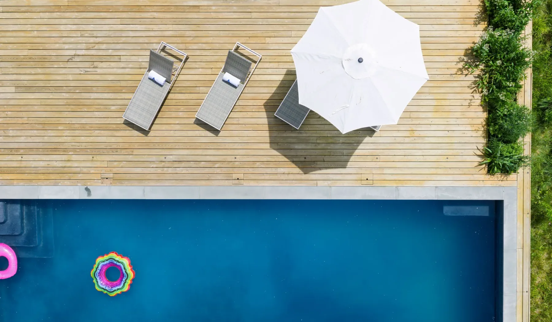 Una piscina junto a una terraza de madera con asientos reclinables y una sombrilla blanca.