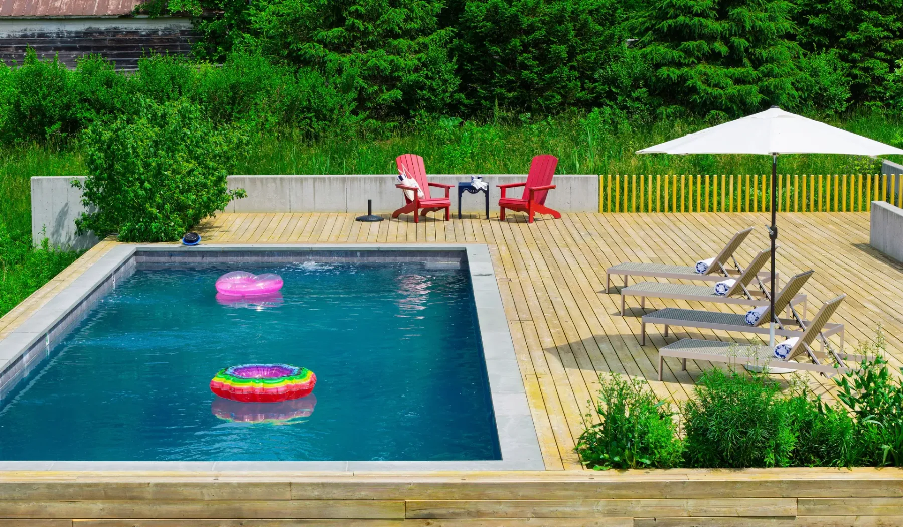 Una piscina al aire libre junto a una terraza de madera con tumbonas y una sombrilla.