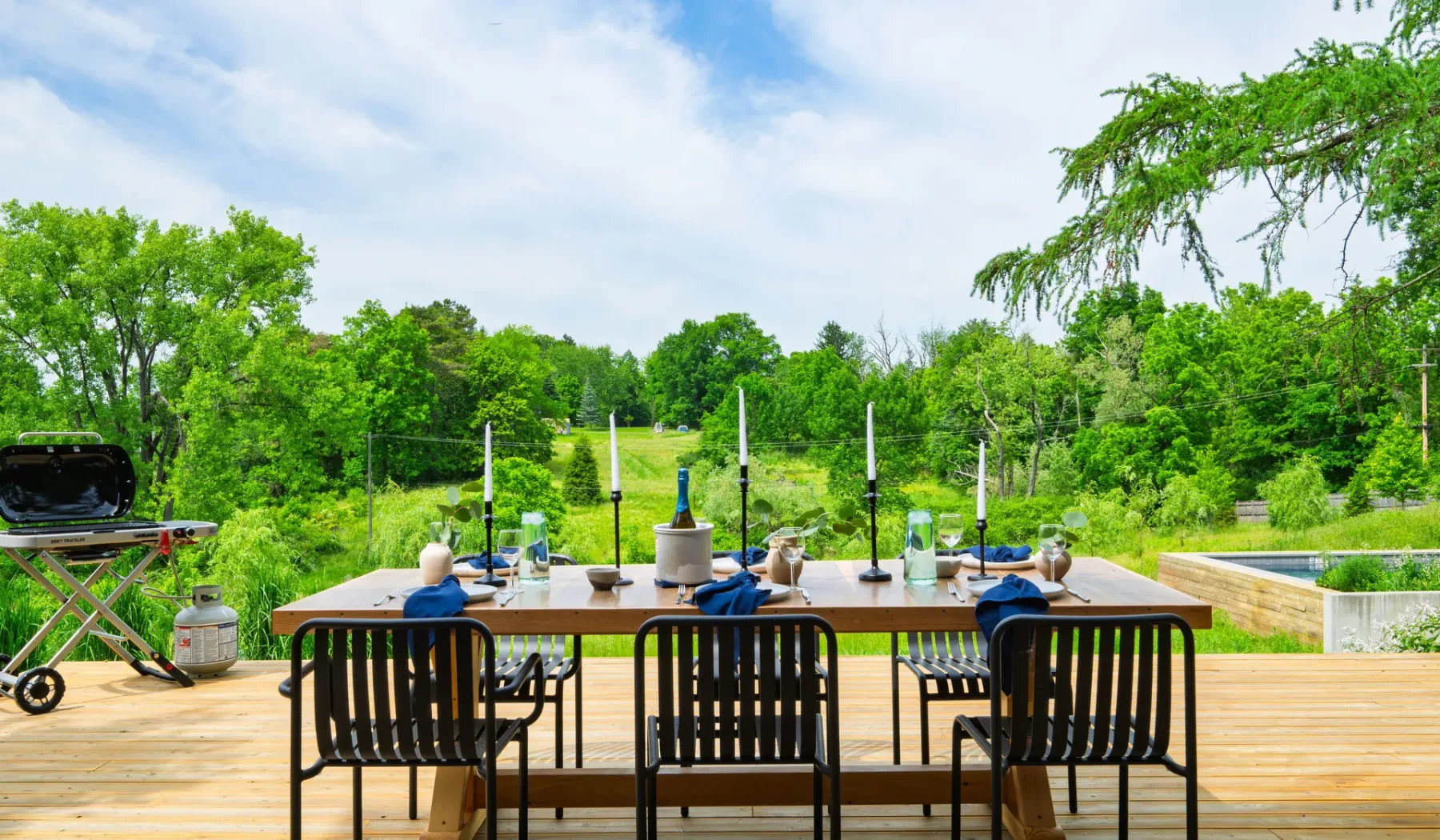 Una mesa de comedor al aire libre con sillas está en una terraza de madera frente a árboles verdes.