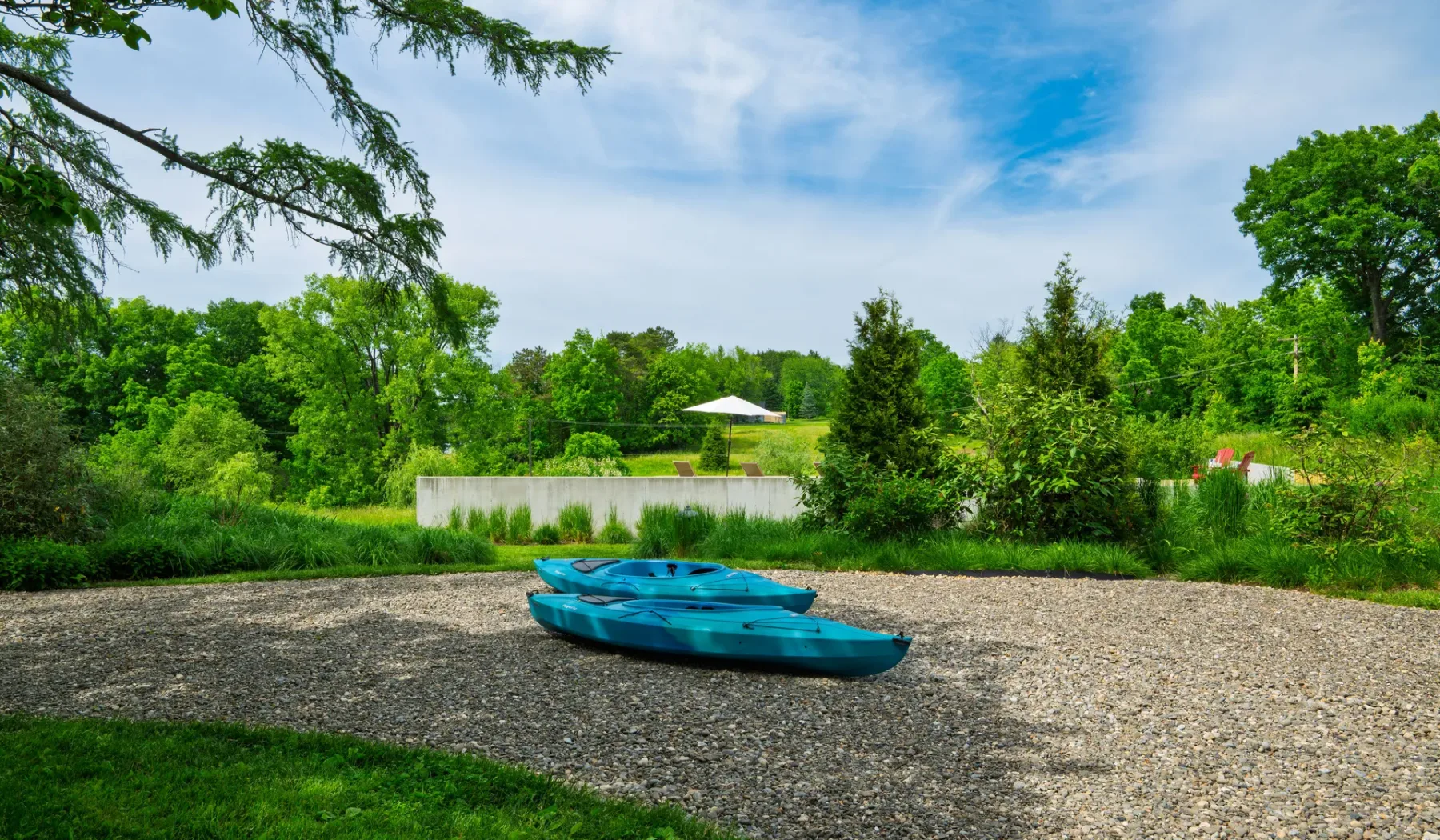 Una orilla de grava con dos kayaks azules, árboles y una pequeña sombrilla blanca al fondo.