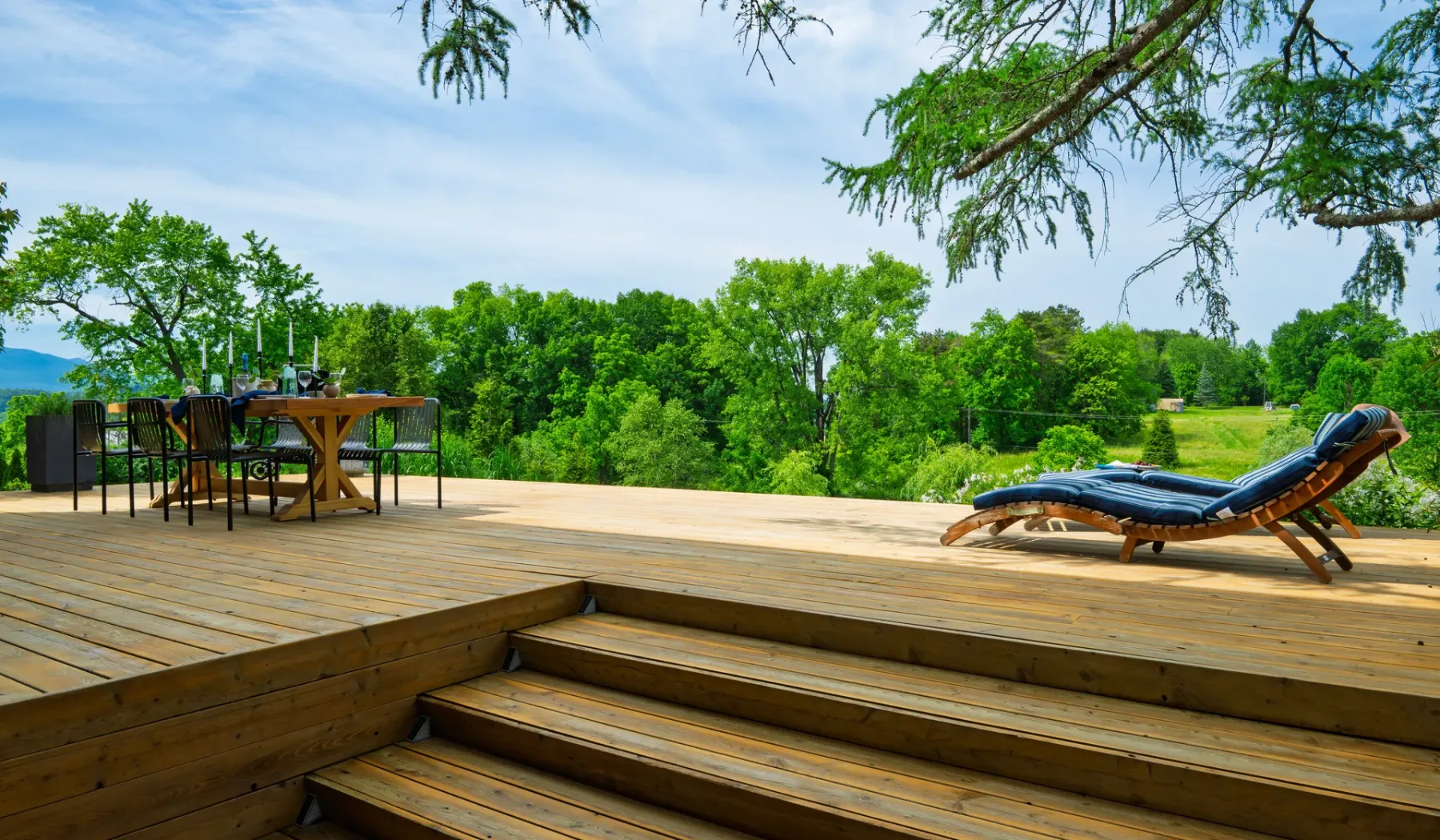 Una terraza de madera con asientos al aire libre y una tumbona da a un paisaje verde.