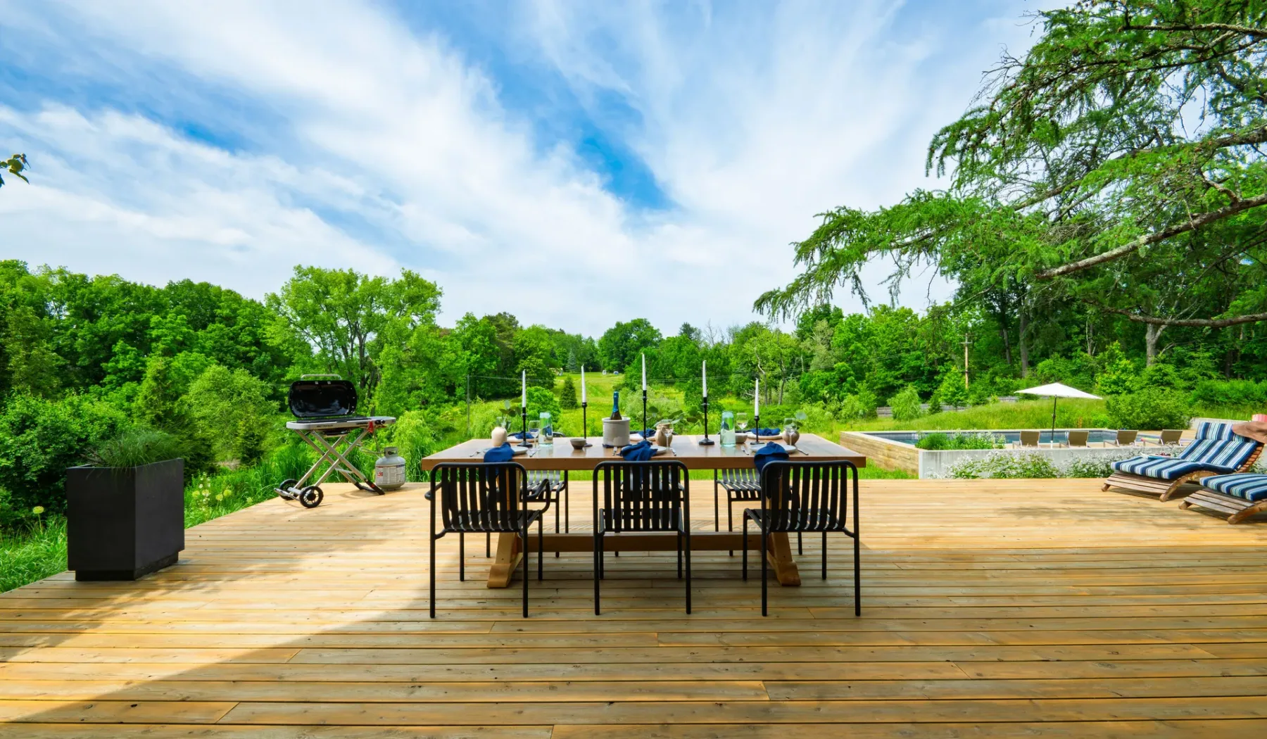 Una mesa y asientos al aire libre están en una amplia terraza de madera junto a una piscina.