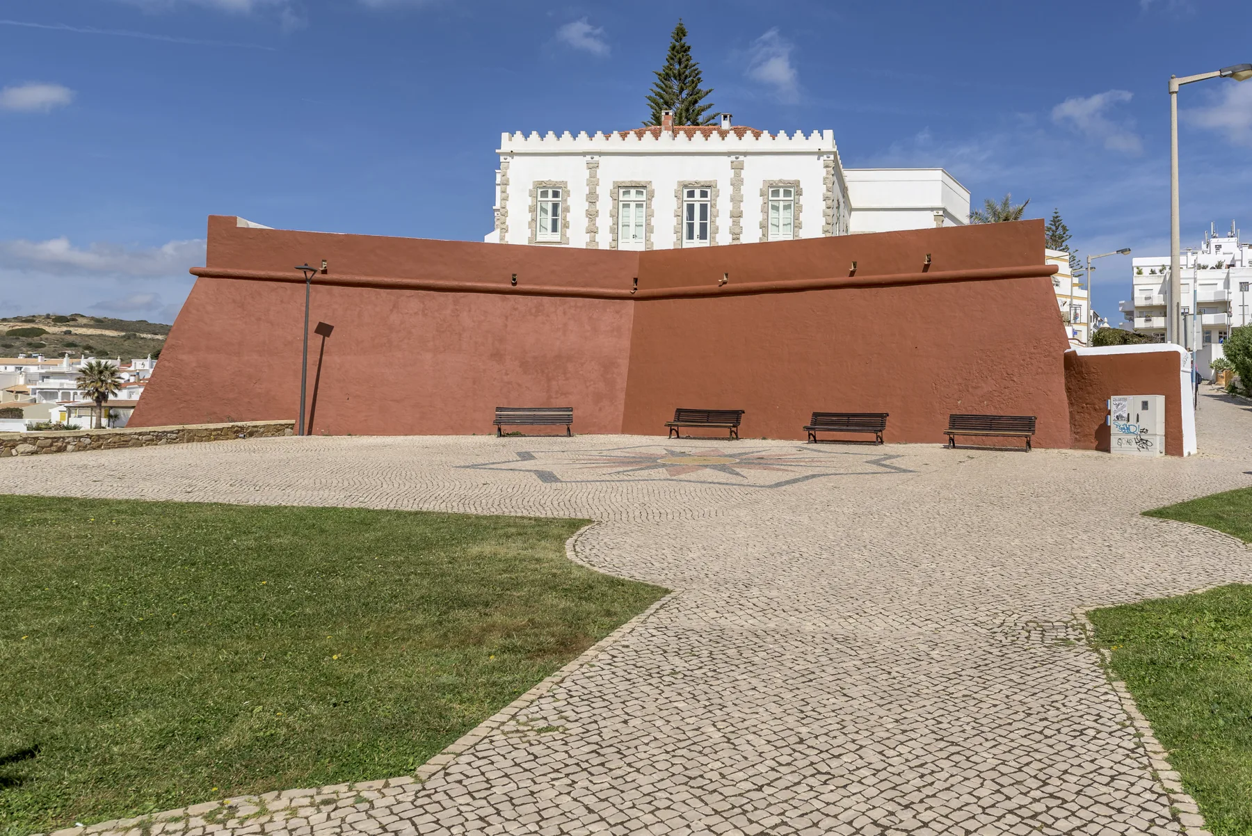 Una plaza pavimentada con bancos se encuentra frente a un muro de color terracota.