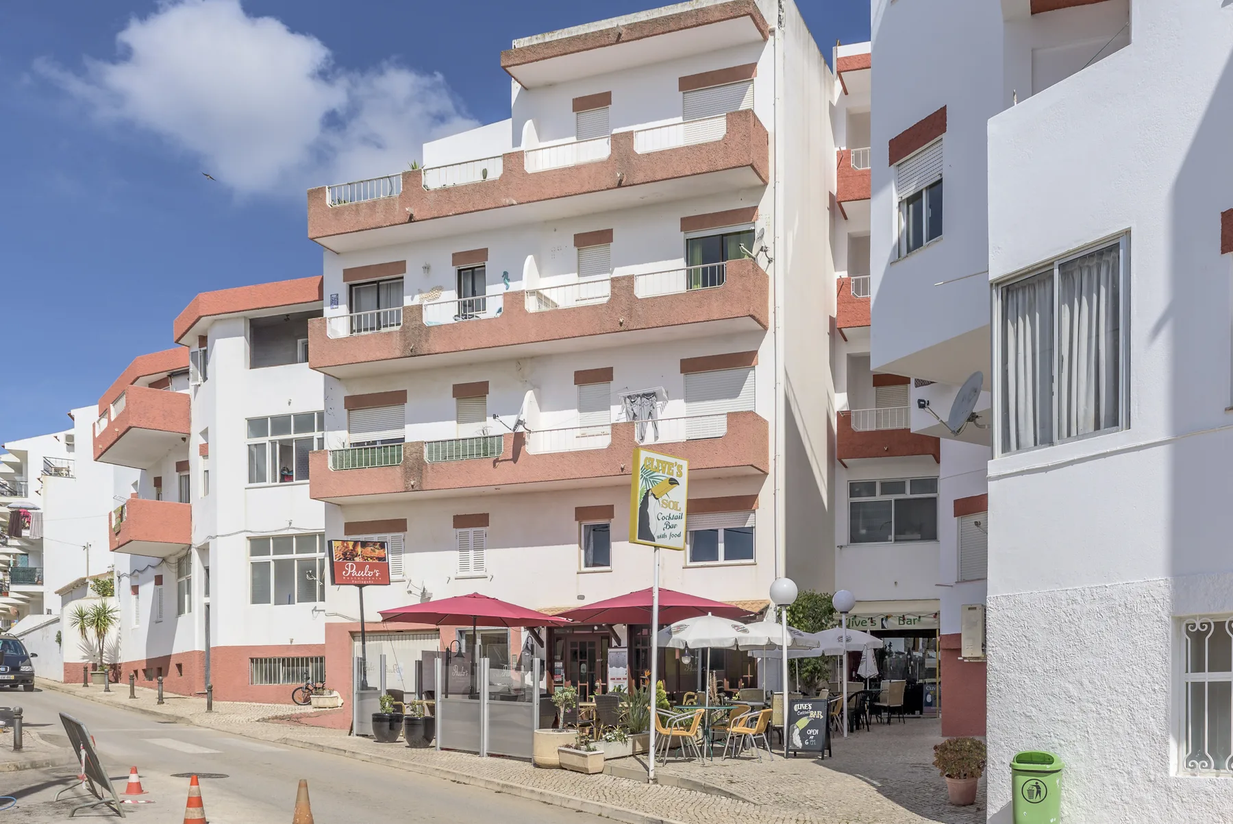 Vista de una calle con un edificio de varios pisos, balcones y mesas de café al aire libre.