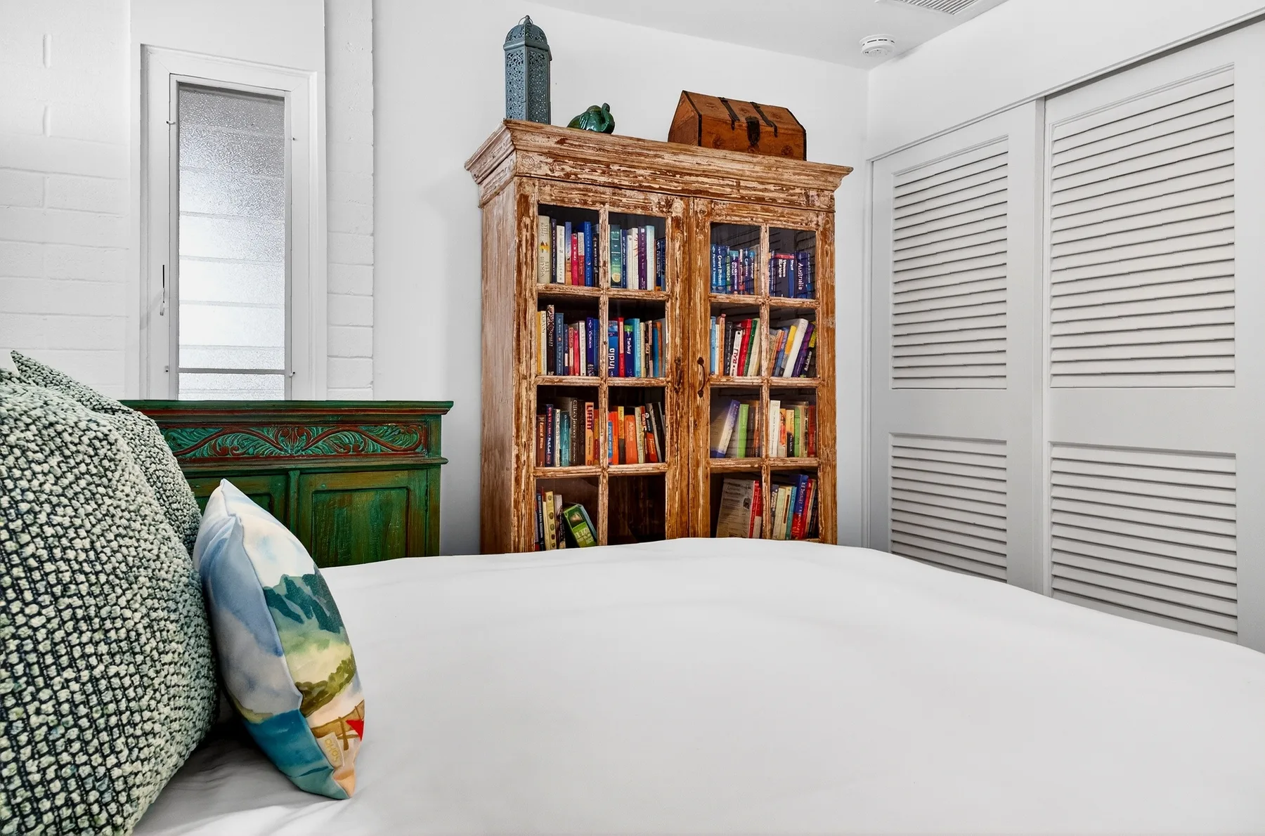 Dormitorio con una cama blanca, cojines estampados y una estantería de madera.