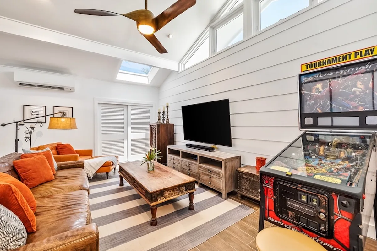 Una sala luminosa con zona de estar, televisor y máquina de pinball.